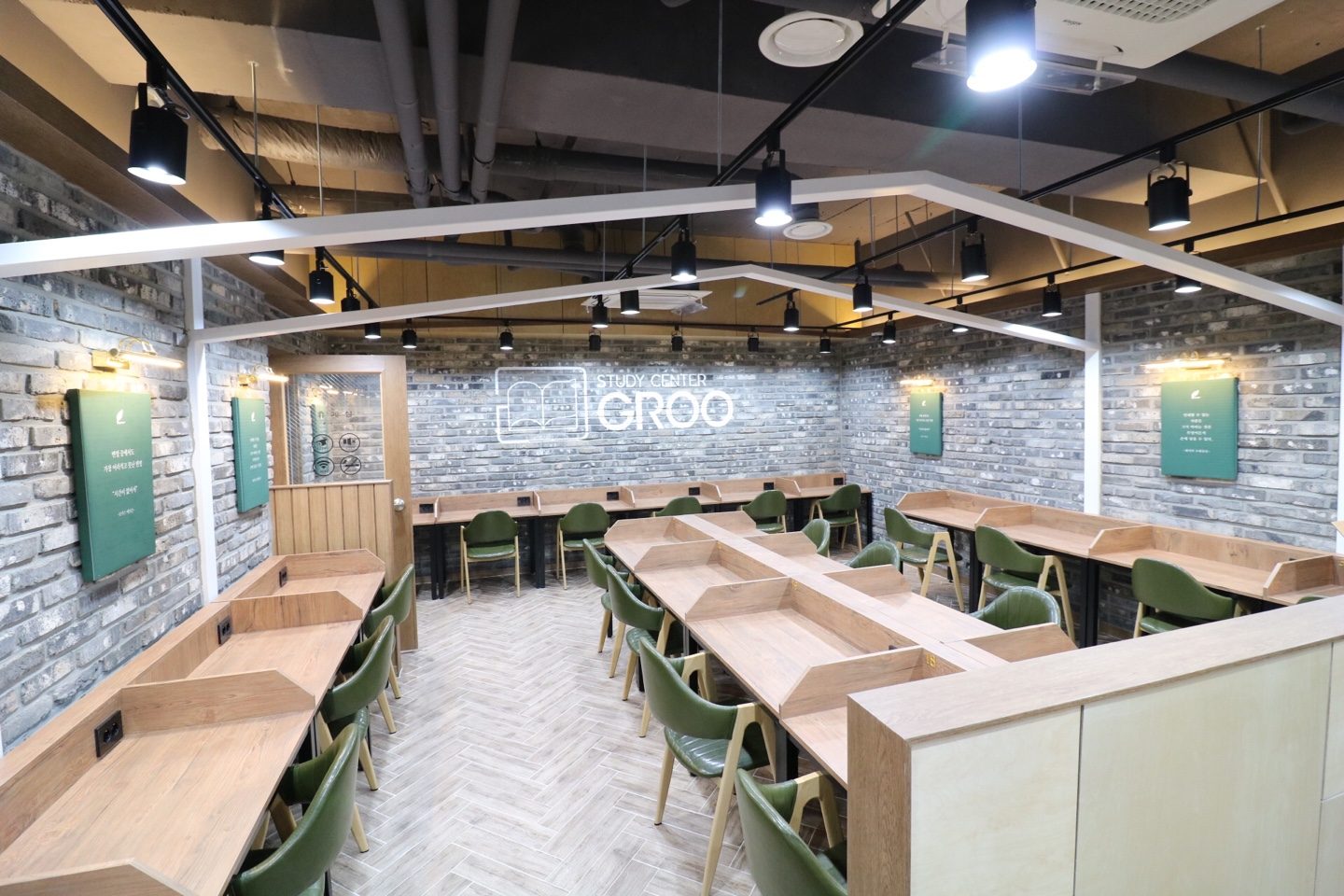 회색 벽돌 벽에 GROO 로고가 있고 나무색 책상과 초록 의자가 놓인 스터디카페 열람실