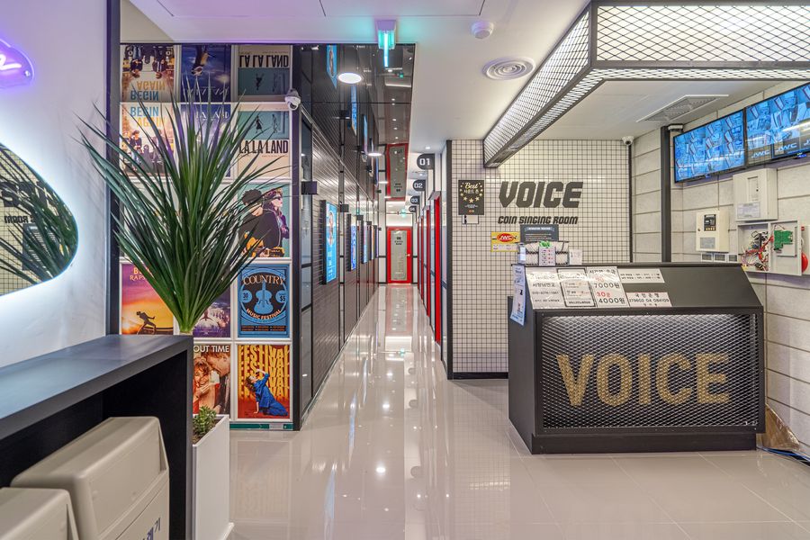VOICE 글자가 박힌 검은 카운터와 화분 뒤로 빨간 문틀의 방들이 늘어선 밝은 코인노래방 복도